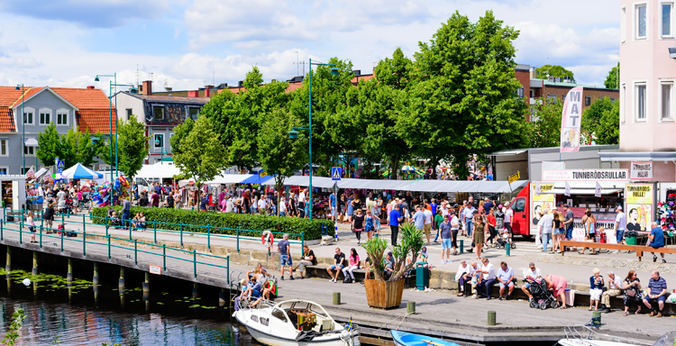 Människor på en brygga och kajpromenad vid ett litet torg en solig dag